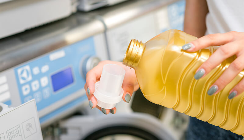 A close-up of a person carefully pouring blue liquid detergent into a white measuring cap, demonstrating the correct way to measure soap for a high-efficiency washing machine.