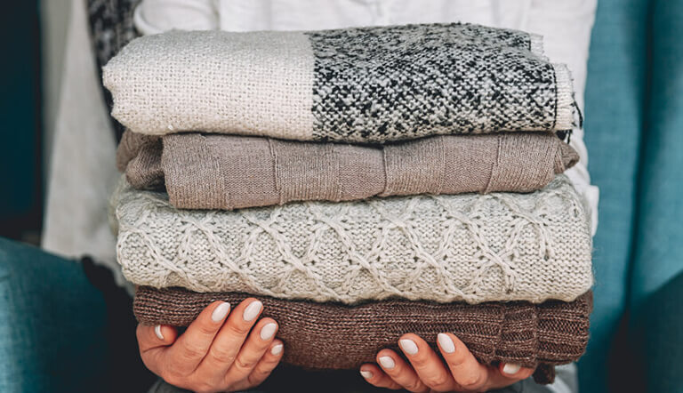 A comparison showing sweaters hung for professional sweater dry cleaning alongside folded knits and washing detergent for home washing.