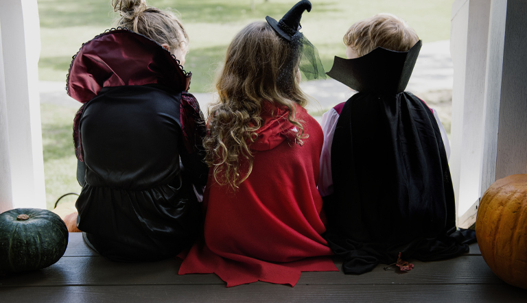 Children wearing Halloween capes and costumes outdoors next to pumpkins.